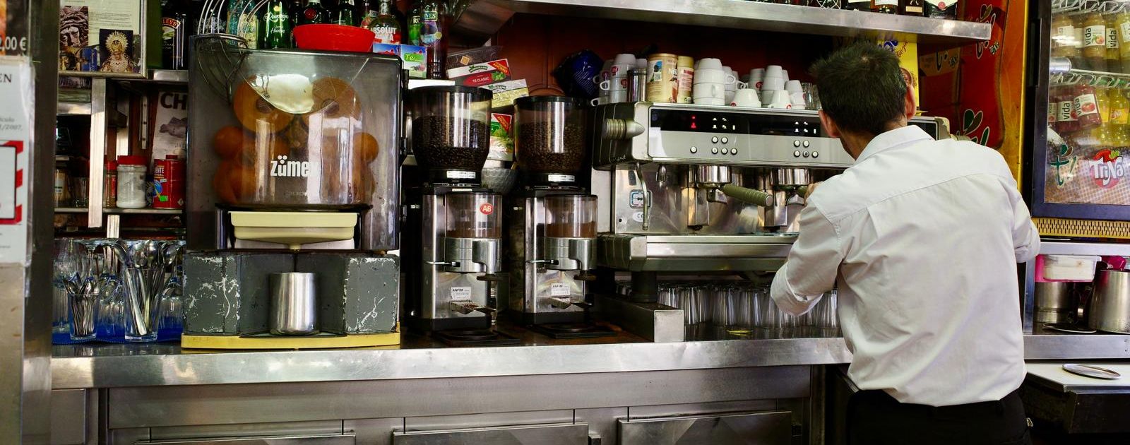 A barman with his back to the camera making coffee at a steel counter in a neighbourhood bar in Spain